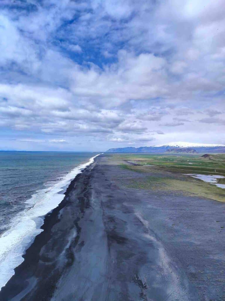 A black sand beach meeting with the waves of the ocean and blue skies behind.