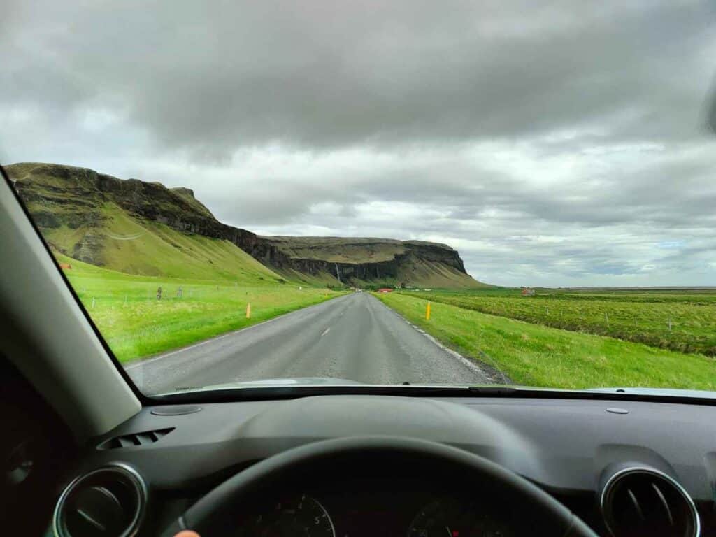 View from a car's front windshield. Driving on an open paved road, with green grass on the sides and mountain ahead.