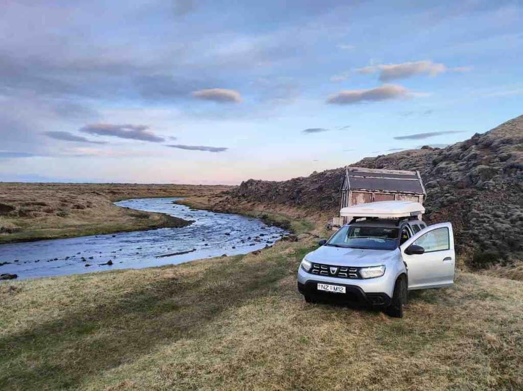 The Duster parked by a small lake. The sun is setting and there is a small tree hut behind.