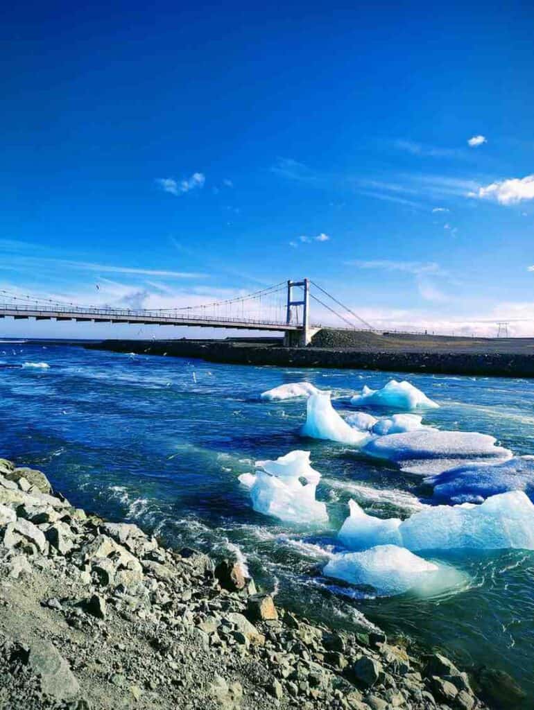 A river with small broken glaciers floating through. Blue skies behind and a bridge in the background.