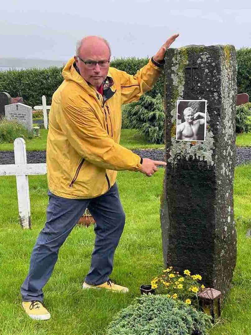 A mean standing by an old graveston with a photo of a muscular man named Jón Páll