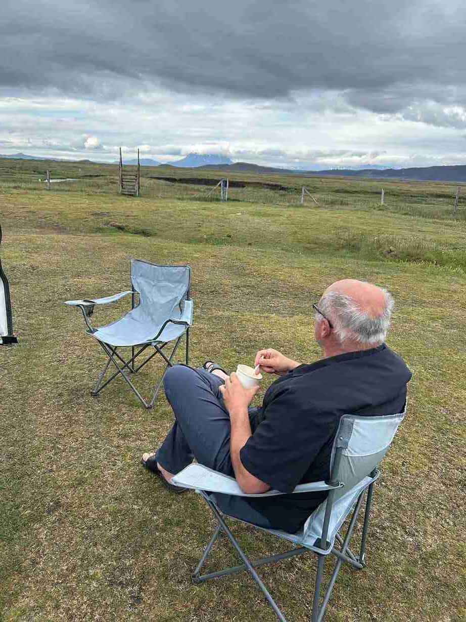 A man sitting in a camping chair in a field, next to another empty camping chair
